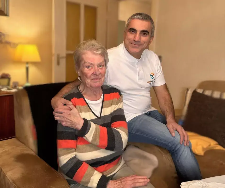 Herzlicher Moment bei Mankas Pflege: Ein Mitarbeiter umarmt eine Kundin zu Hause, während beide freundlich in die Kamera lächeln.